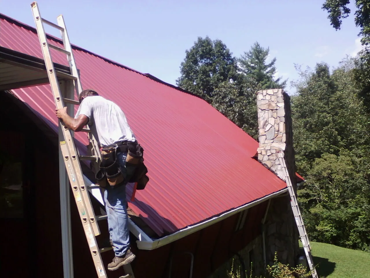 Roofer on a ladder beside a red metal roof and stone chimney — Soffit & Fascia Repair by Roofing Brothers in Crestview